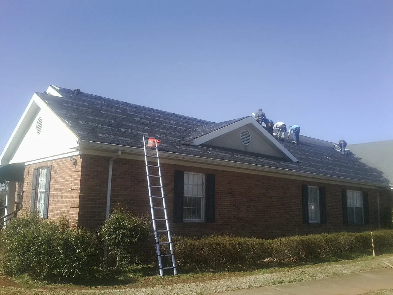 Crew tearing shingles off a brick ranch roof with a ladder set — Metal Roof Repair work by our crew in Clewiston